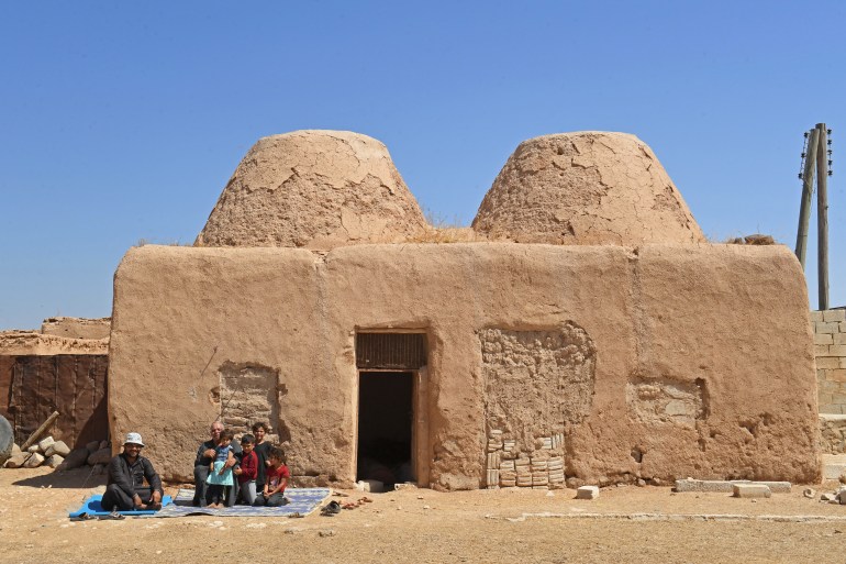 A family sits outside their traditional mud-brick house, known as 'beehive house', in the village of Umm Amuda al-Kabira in Aleppo's eastern countryside, north of Damascus on August 11, 2023. - Traditional mud houses that residents of northern Syria have built for thousands of years risk disappearing, as 12 years of war have emptied villages and left the homes crumbling. The village in Aleppo province is among a handful of villages where residents long used to live in small domed houses made of mud bricks and brittle hay. (Photo by AFP)